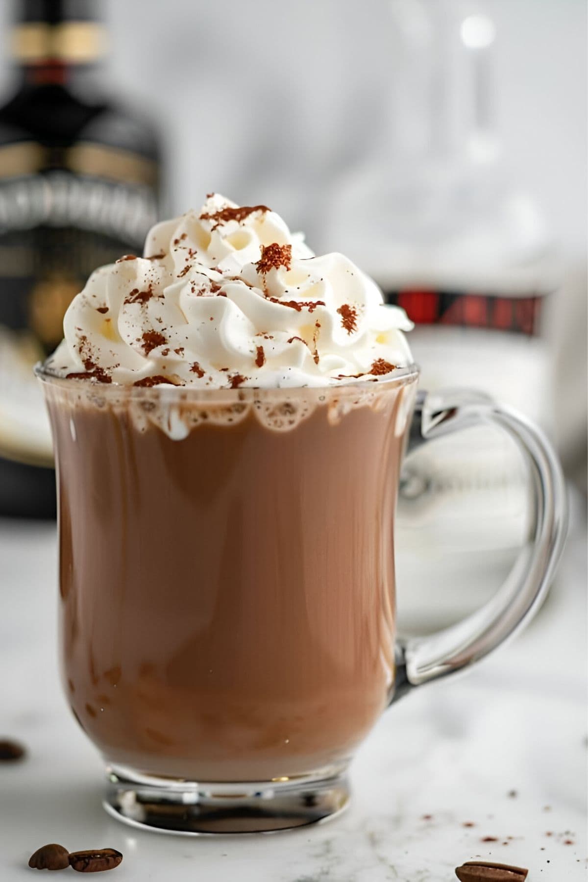 Glass Mug of Bailey's Hot Chocolate with Whipped Cream and Dusted with Cocoa Powder with a Bottle of Bailey's Irish Cream in the Background