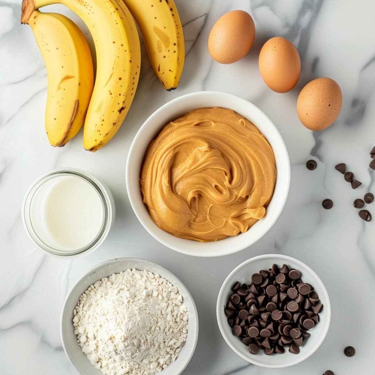 Eggs, bananas, peanut butter, chocolate chips, milk and flour in a white marble table.