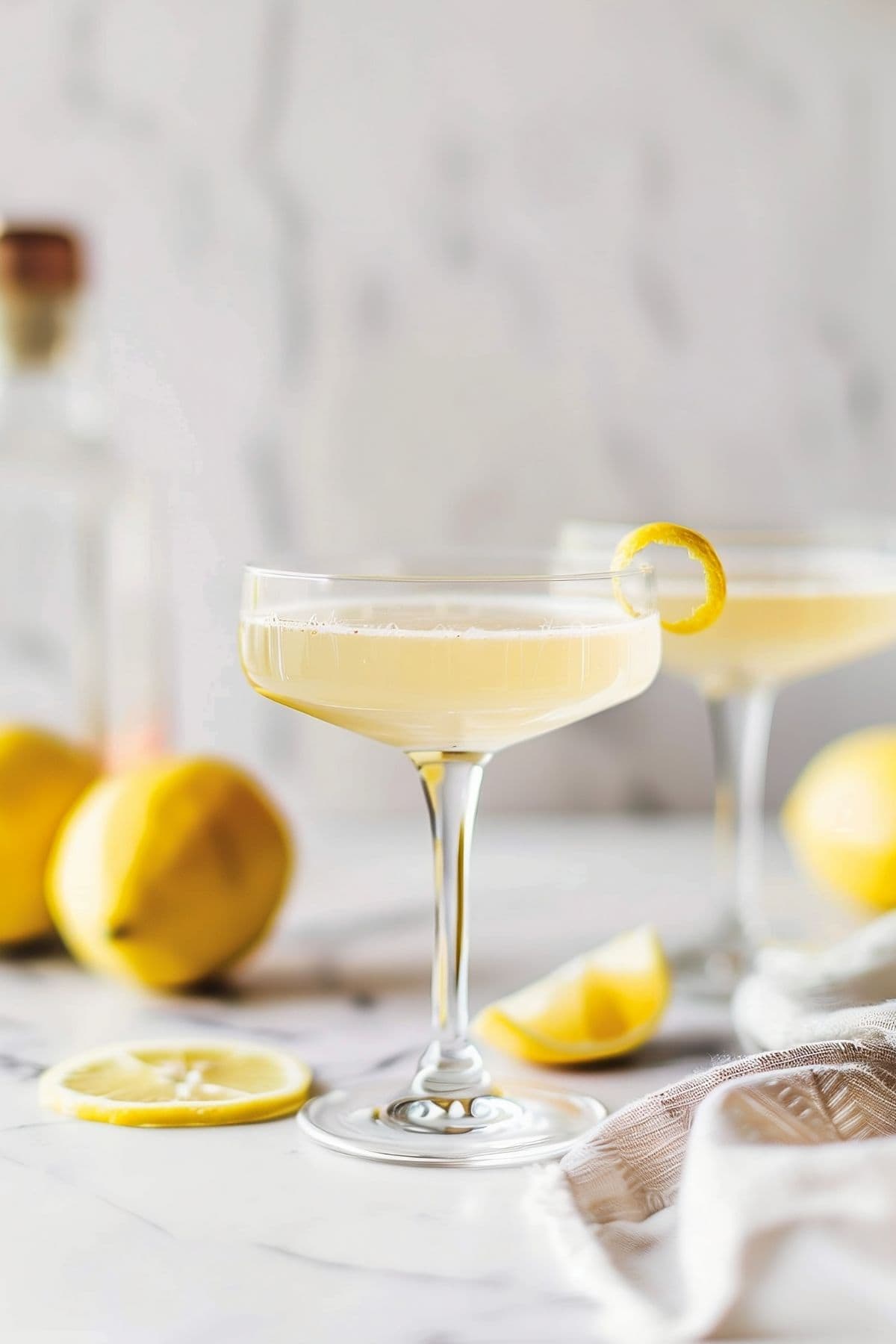Two Bee's Knees Cocktails with a Lemon Peel Twist on a White Marble Table with a Kitchen Towel and Fresh Lemons for Decoration