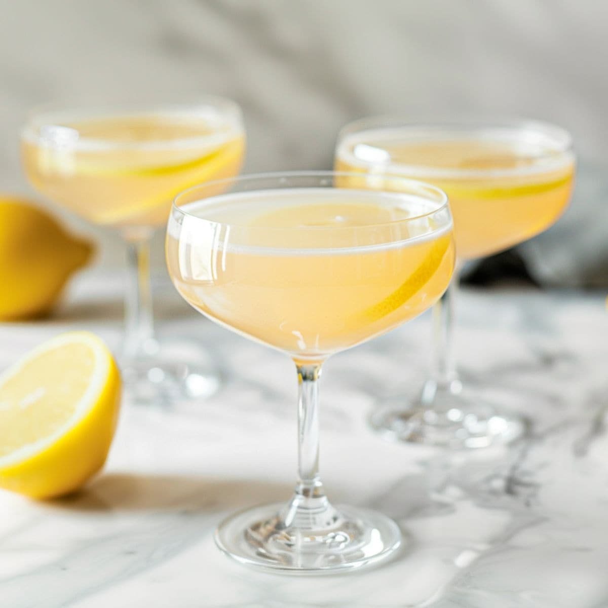Three Bee's Knees Cocktails with Lemon Peel Twists on a White Marble Table with Fresh Lemons 