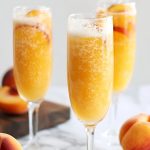 Three Glasses of Bubbly Peach Bellini in Champagne Flutes with Fresh Peach Slices on a White Marble Table with a Wooden Cutting Board and Fresh Peaches All Around