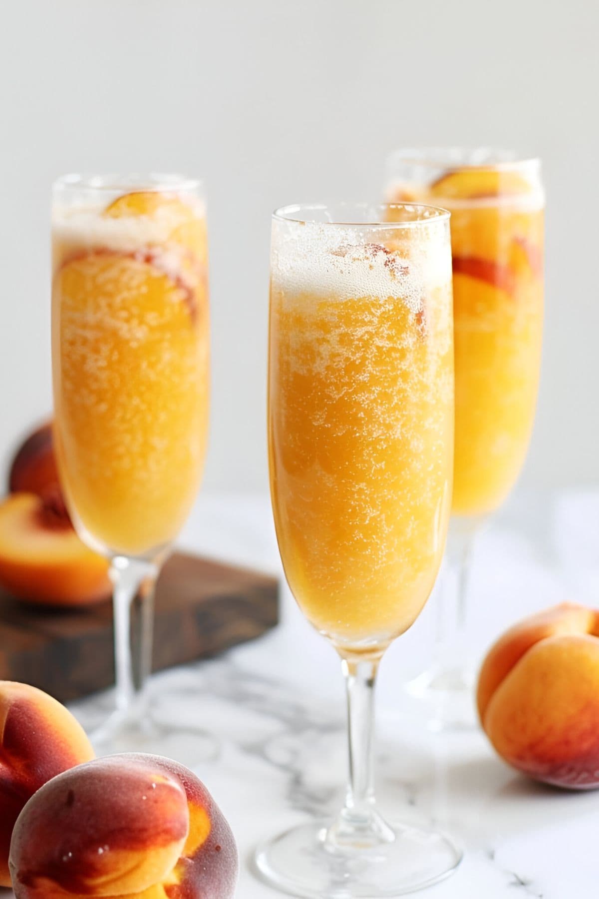 Three Glasses of Bubbly Peach Bellini in Champagne Flutes with Fresh Peach Slices on a White Marble Table with a Wooden Cutting Board and Fresh Peaches All Around
