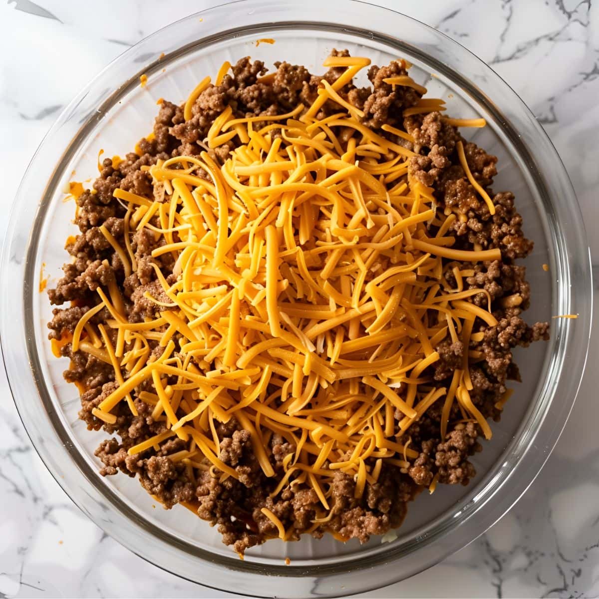 Top View of Ground Beef and Shredded Cheddar Cheese in a Glass Bowl on a White Marble Table