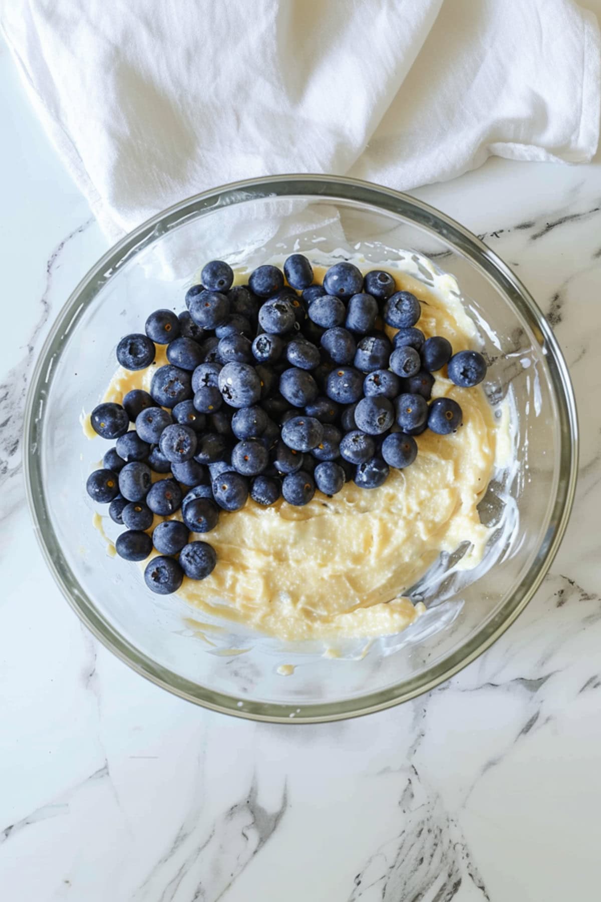 A glass bowl of blueberries and batter on a white marble countertop.