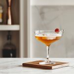 Side View of Brooklyn Cocktail in a Coupe Glass with a Luxardo Maraschino Cherry on a Wooden Coaster on a White Marble Table with a Bar Shelf in the Background