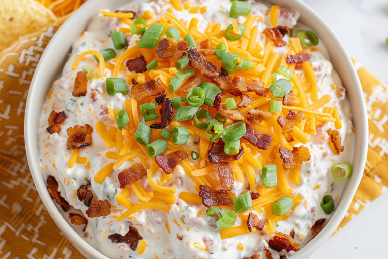 A bowl of crack dip garnished with shredded cheddar cheese, bacon bits and green onions.