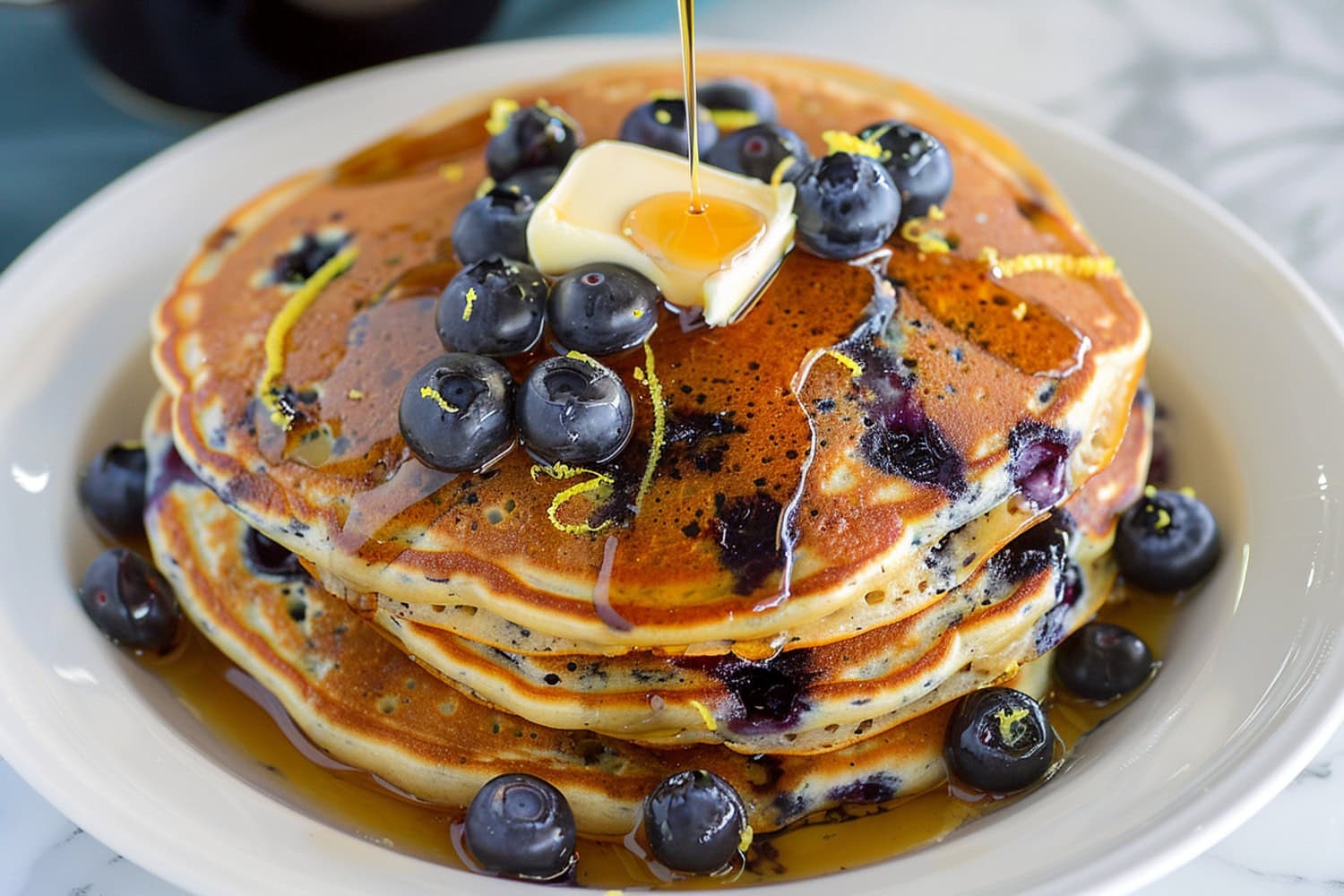Comforting lemon blueberry pancakes, stacked on a plate and served with a generous pour of warm maple syrup.