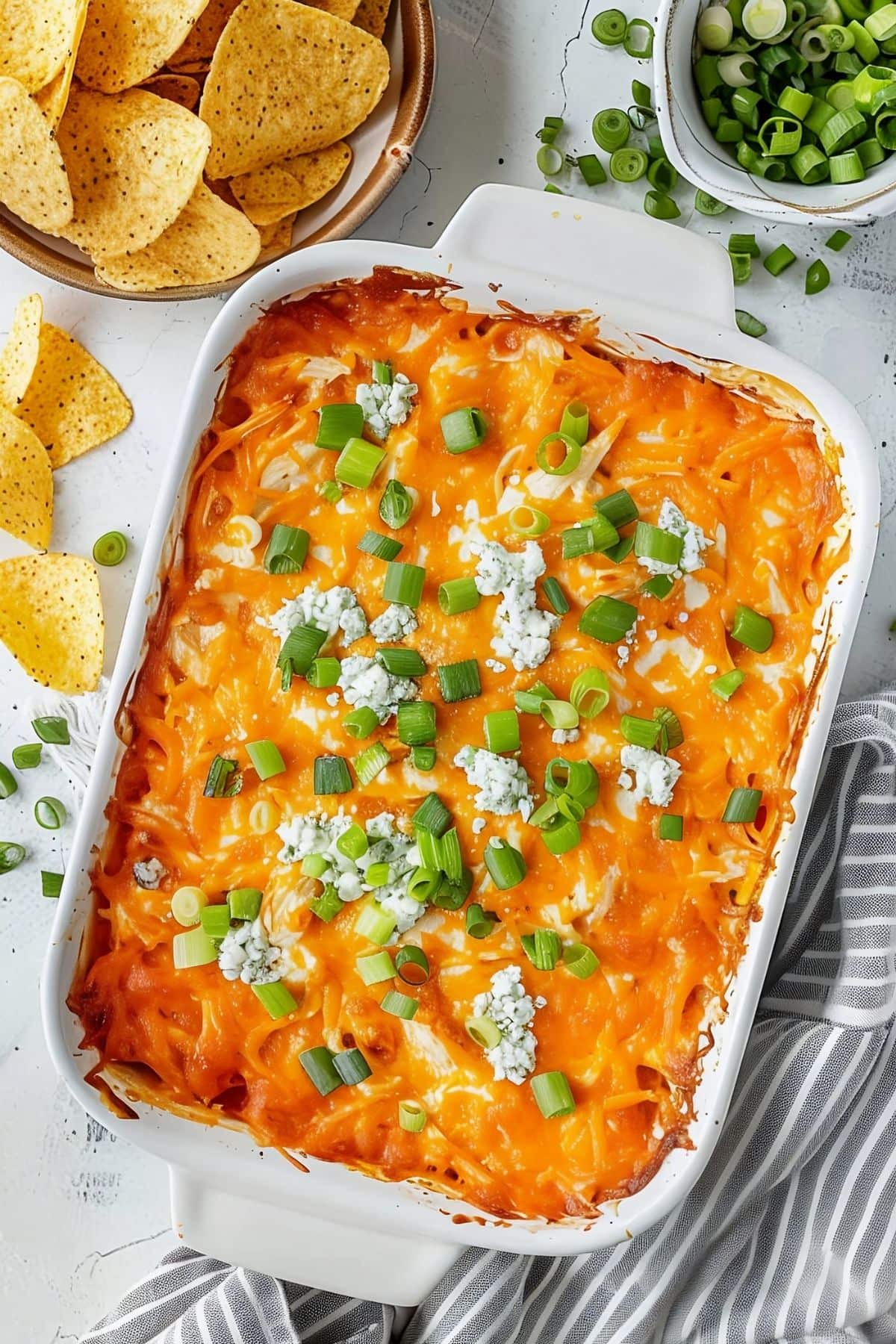 Top View of Frank's Buffalo Chicken Dip Covered with Cheese, Bleu Cheese, and Green Onions in a White Casserole Dish on a White Marble Table with More Green Onions in a Bowl and Tortilla Chips