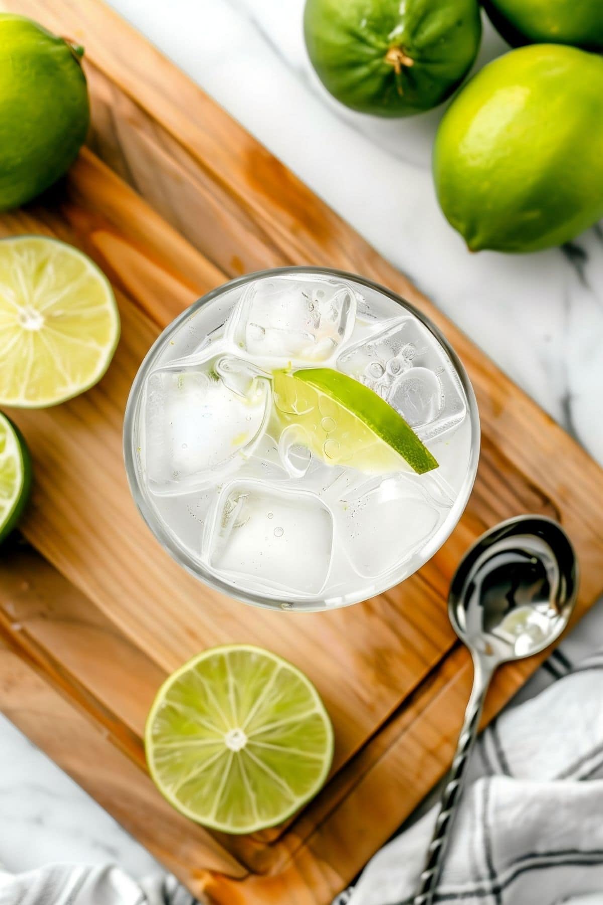 Close Up Top View of Gin and Tonic in a Glass with Ice and a Lime Wedge on a Wooden Cutting Board on a White Marble Table with a Silver Spoon and Limes