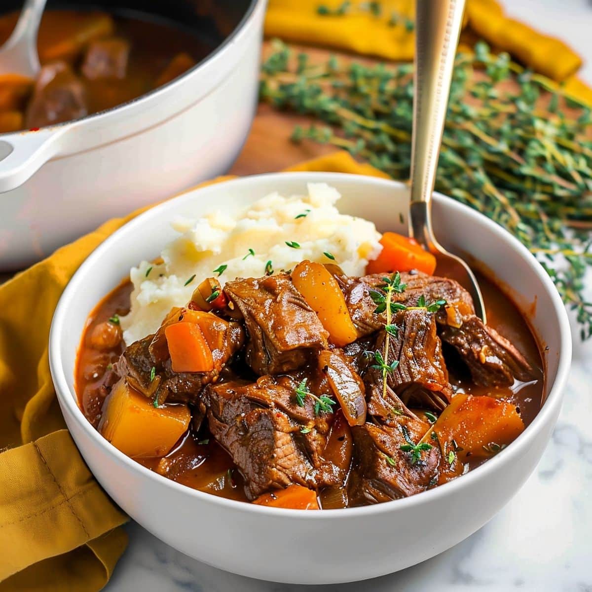 Guinness Beef Stew with Mashed Potatoes in a Bowl Beside a White Dutch Oven and Fresh Herbs to the Side