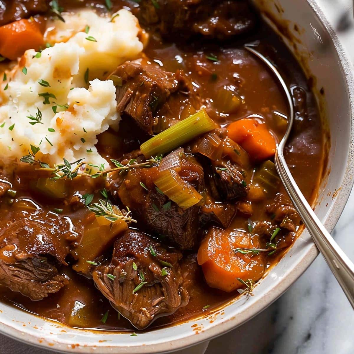 Guinness Beef Stew in a White Bowl with Meaty Beef, Celery, and Carrots over Mashed Potatoes with a Spoon