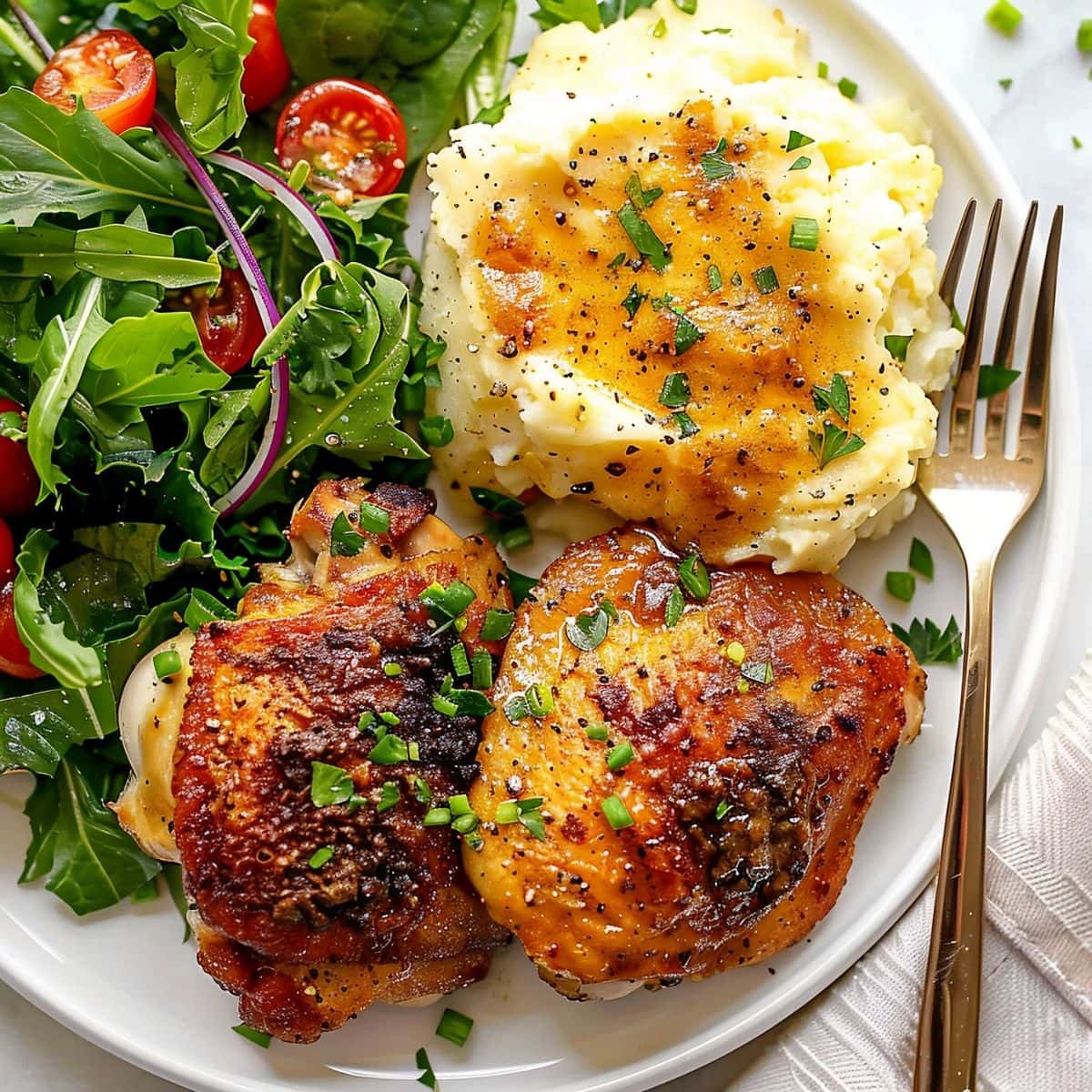 Close Top View of Two Crispy Baked Chicken Thighs with Mashed Potatoes, Gravy, and a Side Salad on a White Plate with a Fork on a White Marble Table