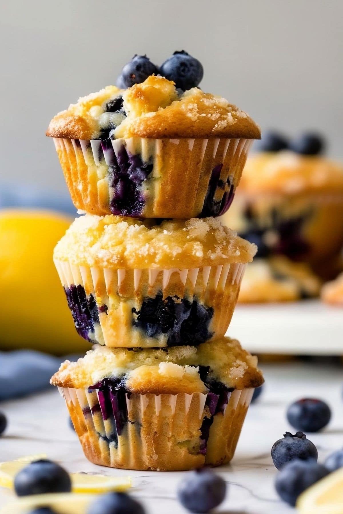 Three Stacked Lemon Blueberry Muffins on a White Marble Table with Blueberries Around the Muffins
