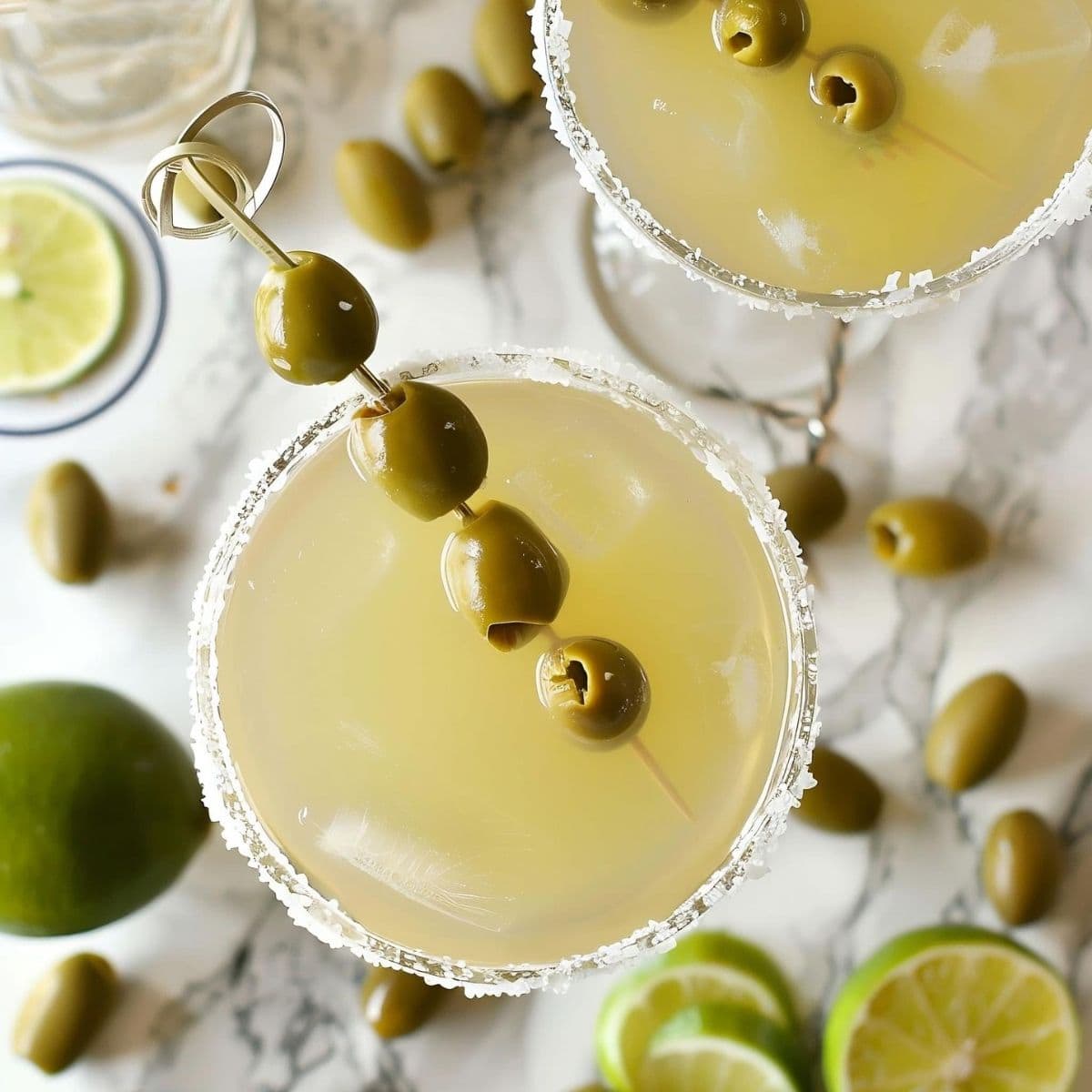 Top View Mexican Martinis with Salted Rims and Olives on a White Marble Table with Limes and Olives Around the Table