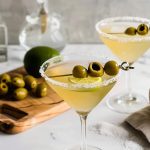 Two Mexican Martinis with Olives on a White Marble Table with a Cutting Board with Lemons and Olives in the Background