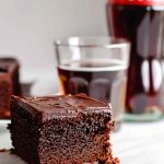 Slice of Chocolate Coca Cola Cake on a Piece of Parchment Paper on a White Marble Table with a Glass of Cola and a Glass Bottle of Coca Cola in the Background