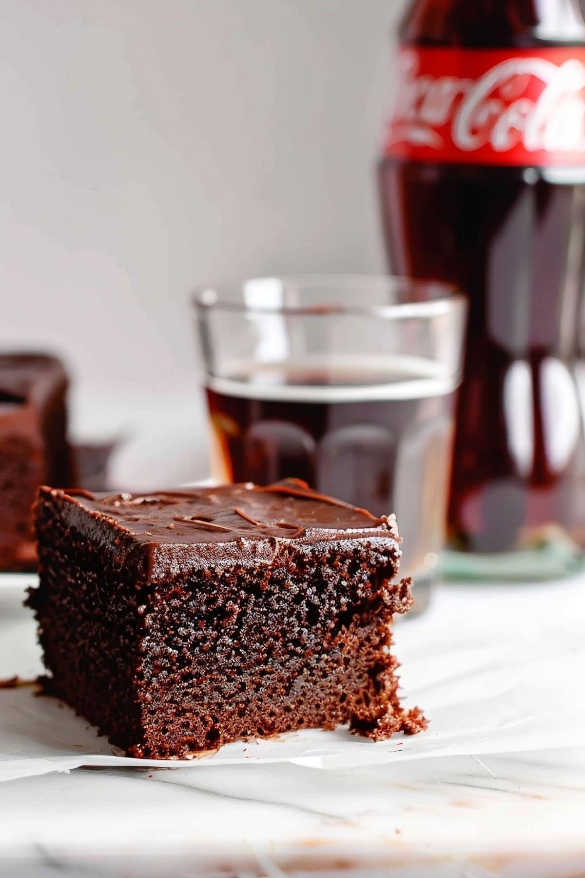 Slice of Chocolate Coca Cola Cake on a Piece of Parchment Paper on a White Marble Table with a Glass of Cola and a Glass Bottle of Coca Cola in the Background