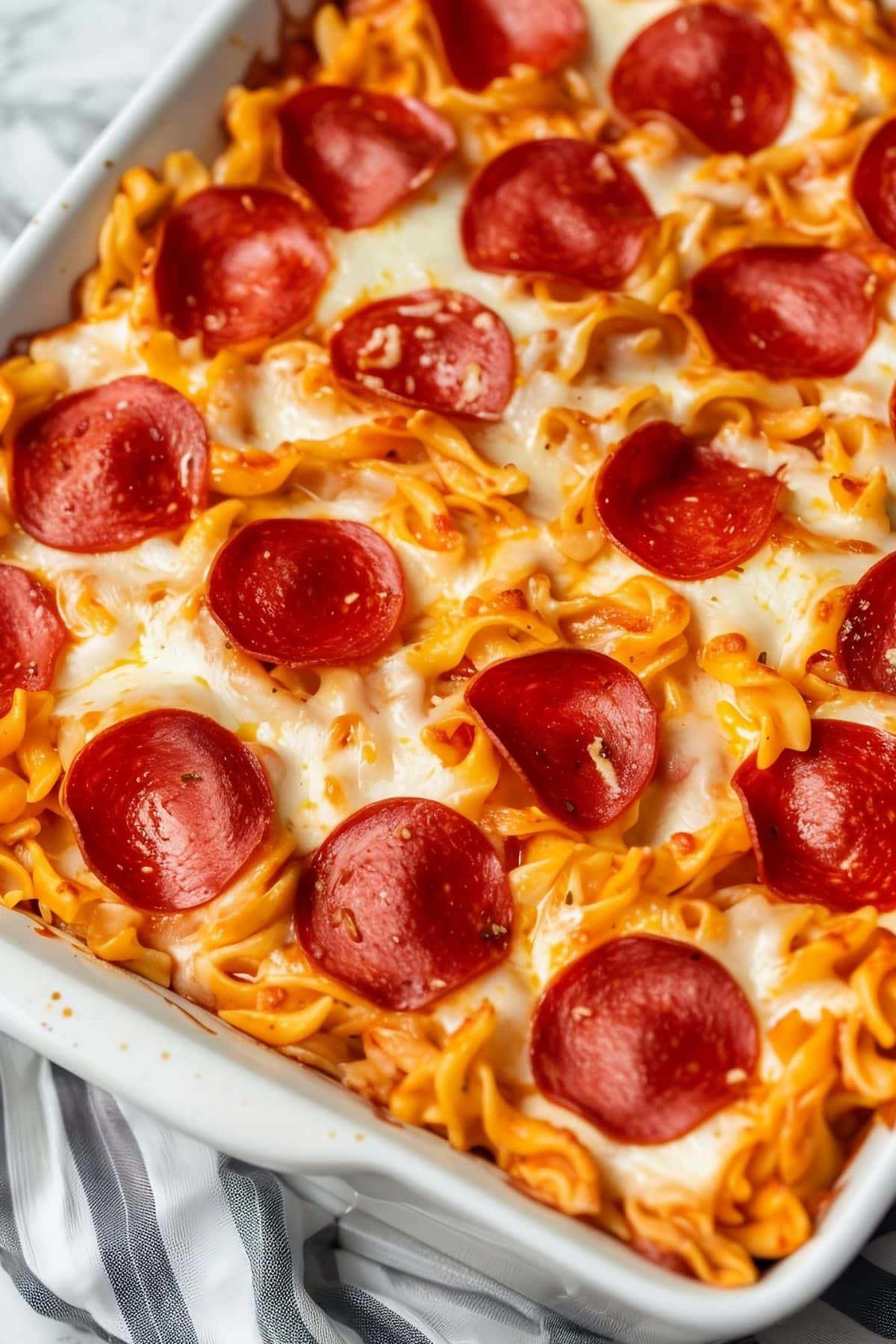 Close Up View of Pepperoni Pizza Casserole in a White Casserole Dish with Egg Noodles, Melted Cheese, and Slices of Pepperoni