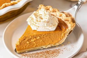 Pie ladle with a slice of pumpkin cream pie on a white plate topped with whipped cream.