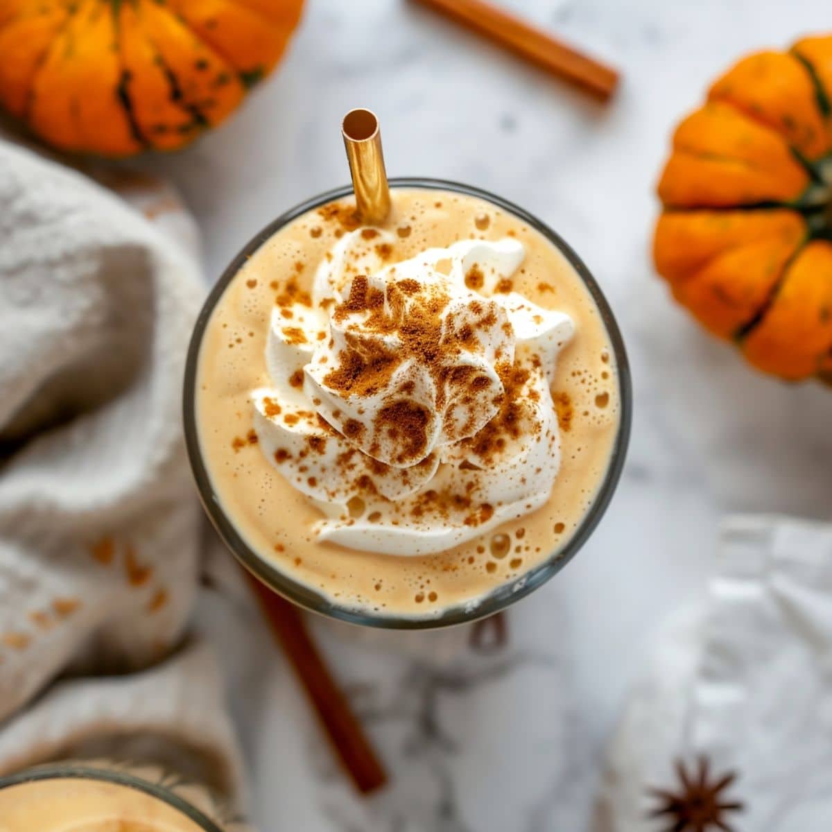 Top View Pumpkin Milkshake in a Glass with Whipped Cream, Cinnamon, and a Straw on a White Marble Table with Pumpkins and Cinnamon Sticks