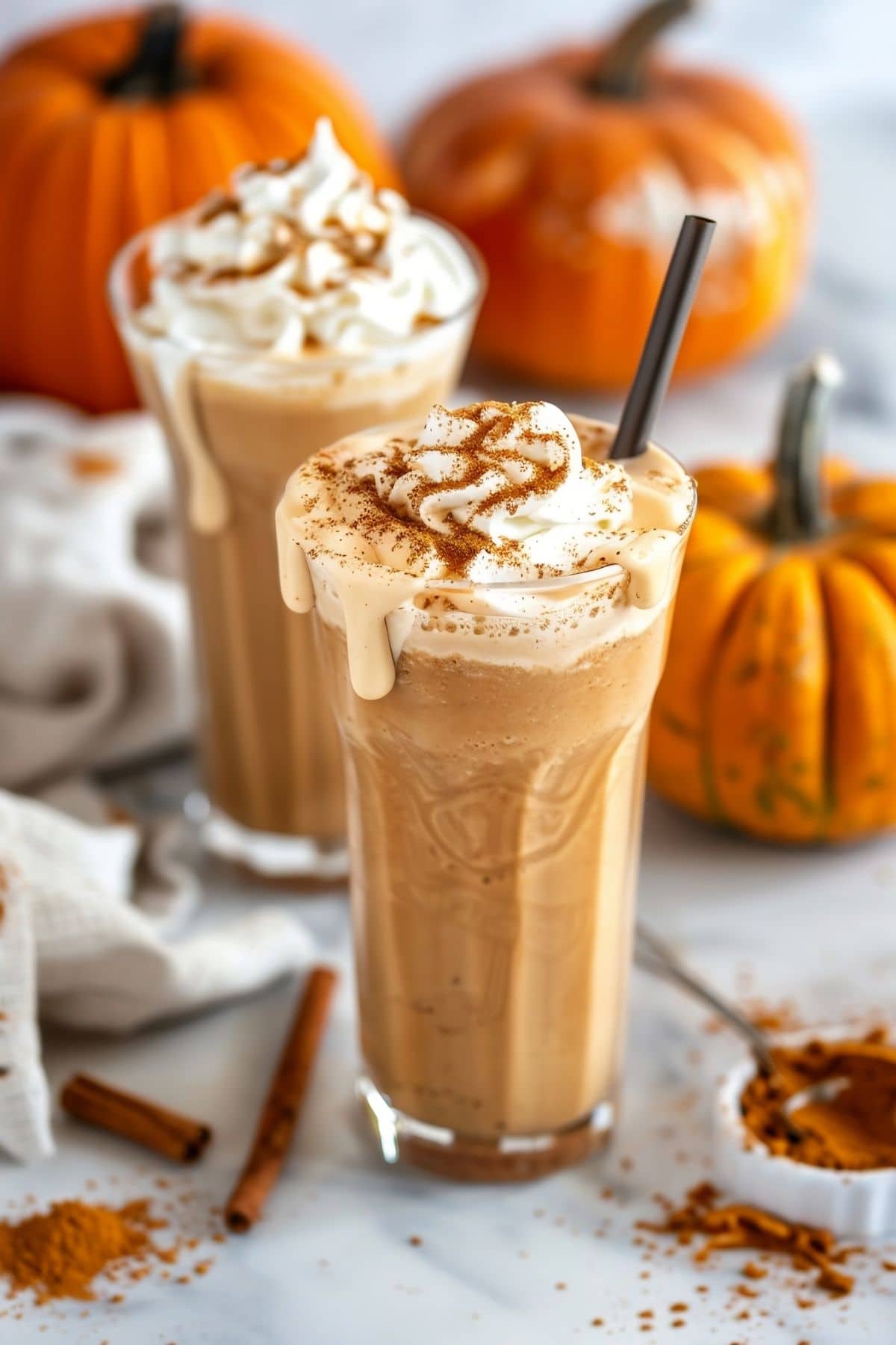 Two Pumpkin Milkshakes in Tall Glasses with Whipped Cream and Cinnamon on a White Marble Table Decorated with Pumpkins and Cinnamon Sticks