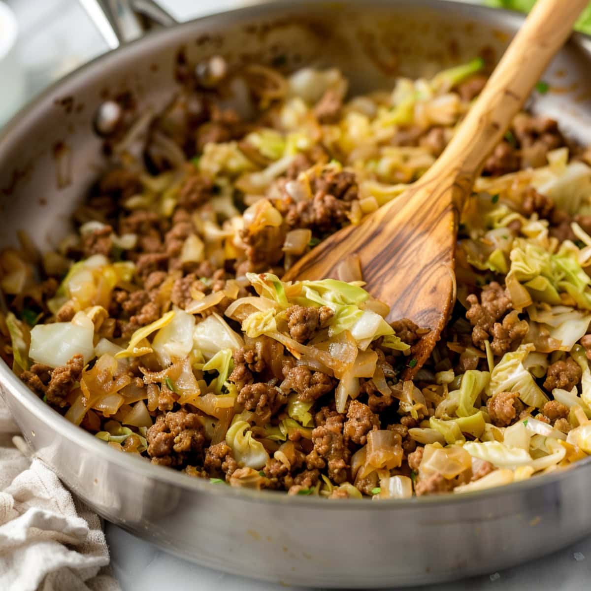 Ground pork and cabbage tossed in a stainless skillet with wooden ladle.