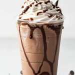 Copycat Starbucks Double Chocolate Chip Frappuccino with Whipped Cream and a Chocolate Drizzle in a Tall Plastic Cup with a Striped Straw and More Chocolate Chips on the Table Around the Cup
