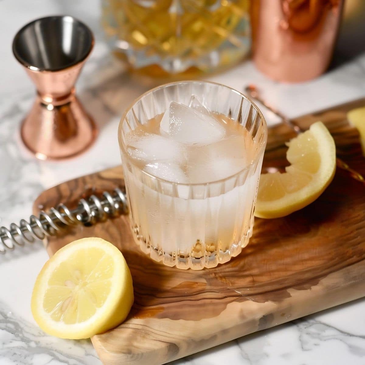 White Negroni in a Rocks Glass with Ice on a Wooden Cutting Board with Cut Lemons and Rose Gold Cocktail-Making Equipment