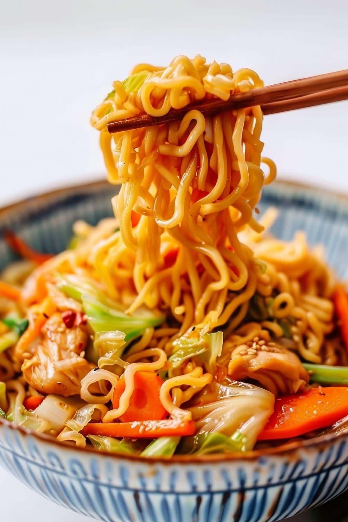 Close Up of Chopsticks Holding Noodles from the Yakisoba Chicken Dish in a Bowl with Chicken, Soba Noodles, and Veggies