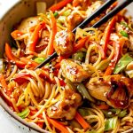Close Up Yakisoba Chicken with Soba Noodles, Meaty Chicken, Peppers, Onions, Carrots, Cabbage, and Sesame Seeds Covered in Sauce in a Bowl with Chopsticks