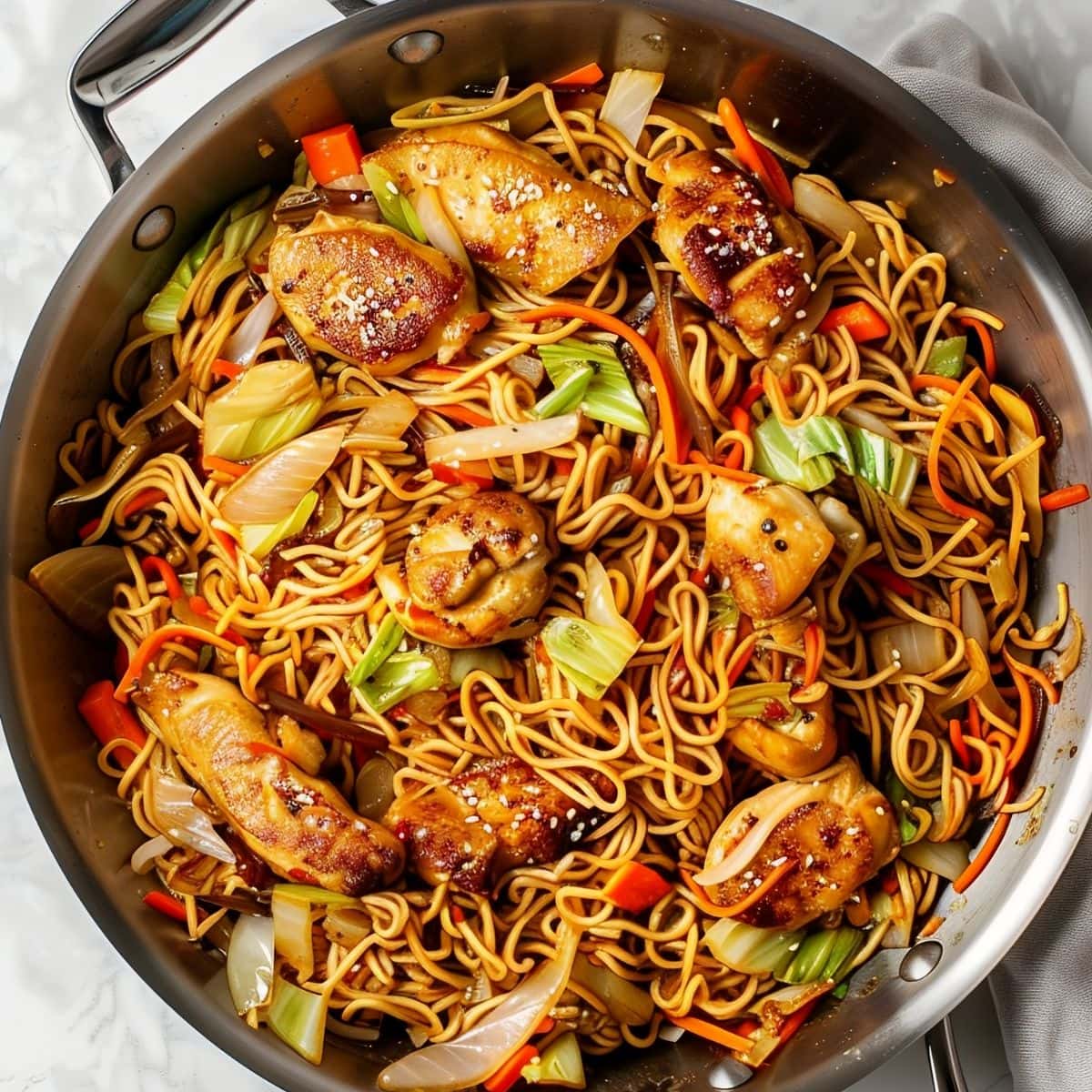 Top View of Yakisoba Chicken in the Frying Pan with Chunks of Chicken, Soba Noodles, Onions, Cabbage, Carrots, and Peppers in Sauce