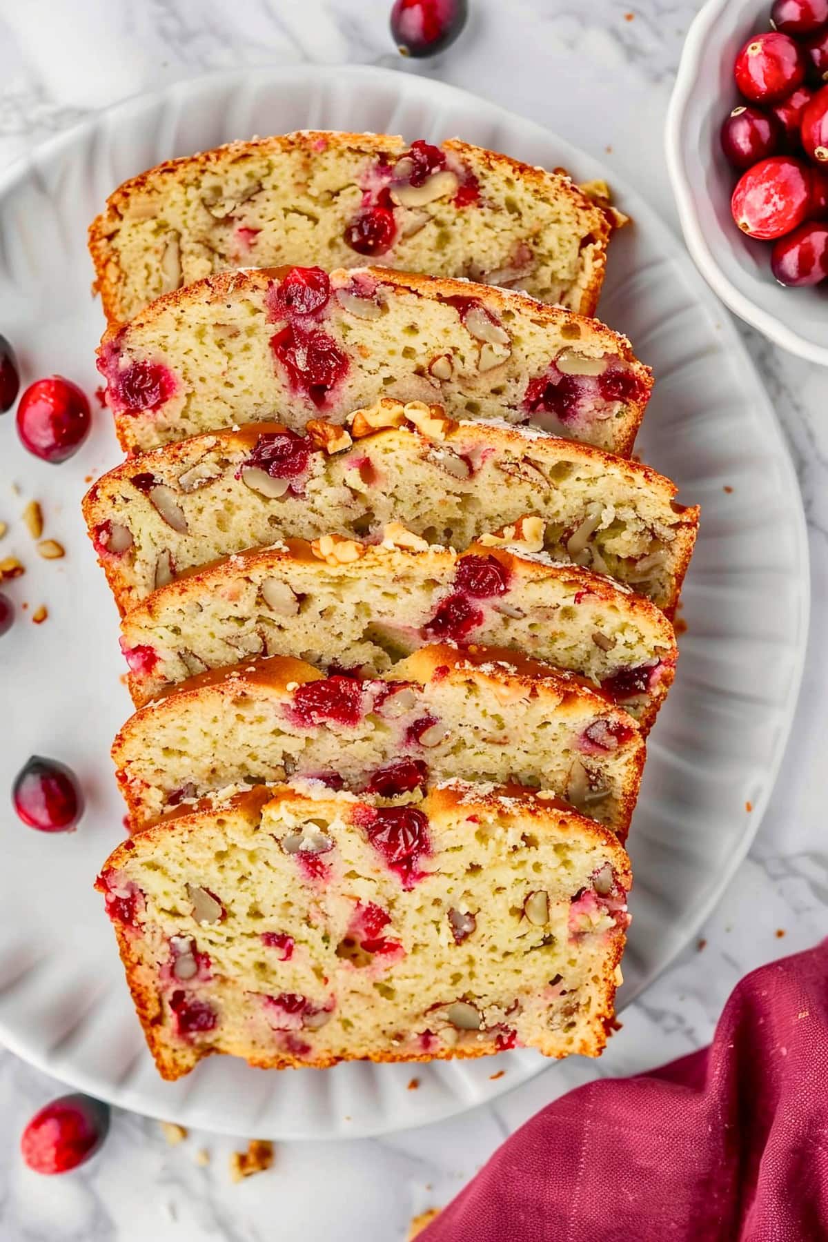 Slices of Cranberry Orange Bread Filled with Walnuts and Cranberries on a White Plate on a White Marble Table with Cranberries All Around