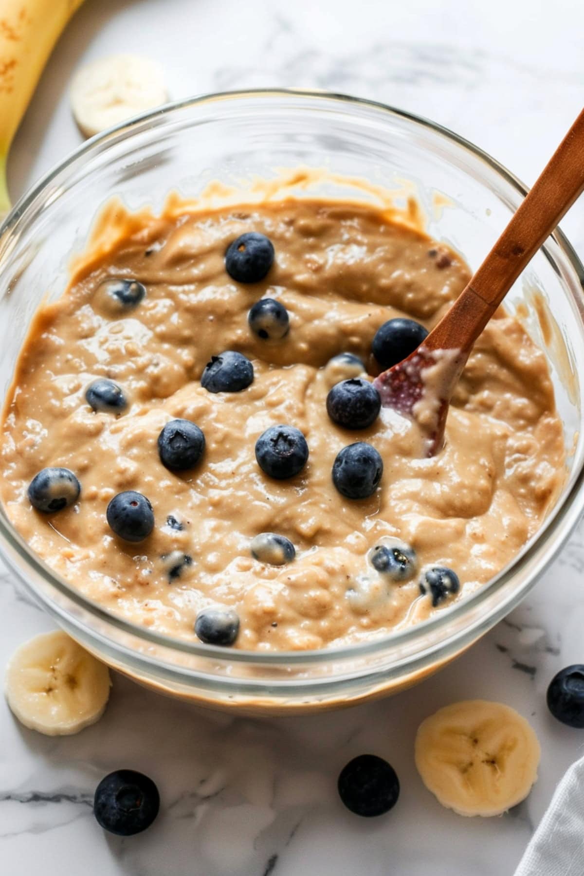 Muffin batter with banana slices and blueberries on a white marble table.