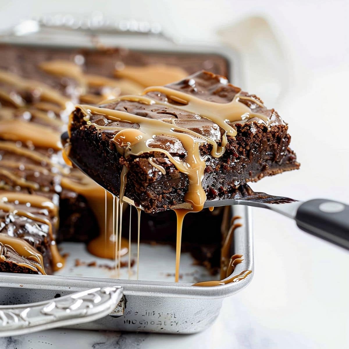 Spatula Holding Up a Caramel Brownie Square Dripping Caramel over a Baking Sheet of More Caramel Brownies