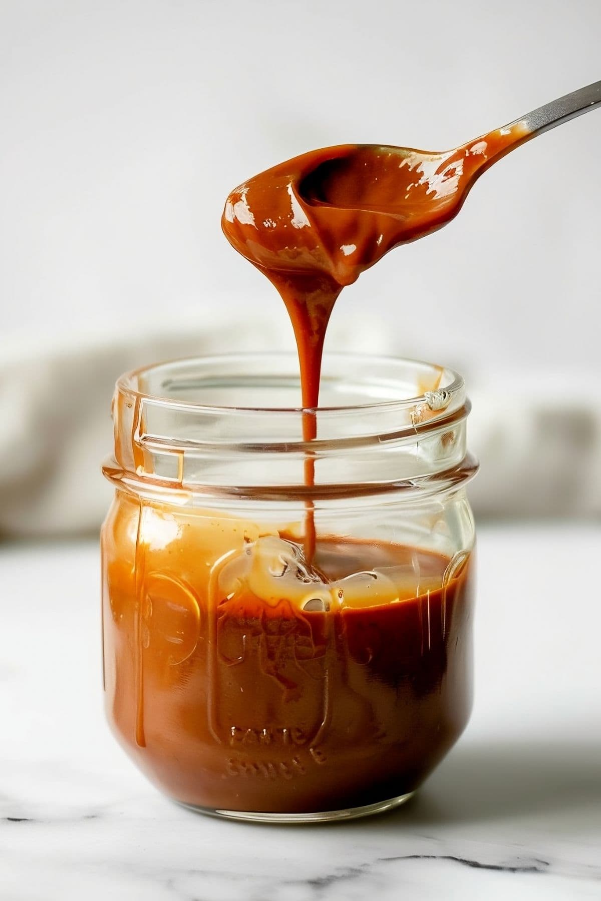 Jar of Salted Caramel with a Spoon Dripping Caramel on a White Marble Table