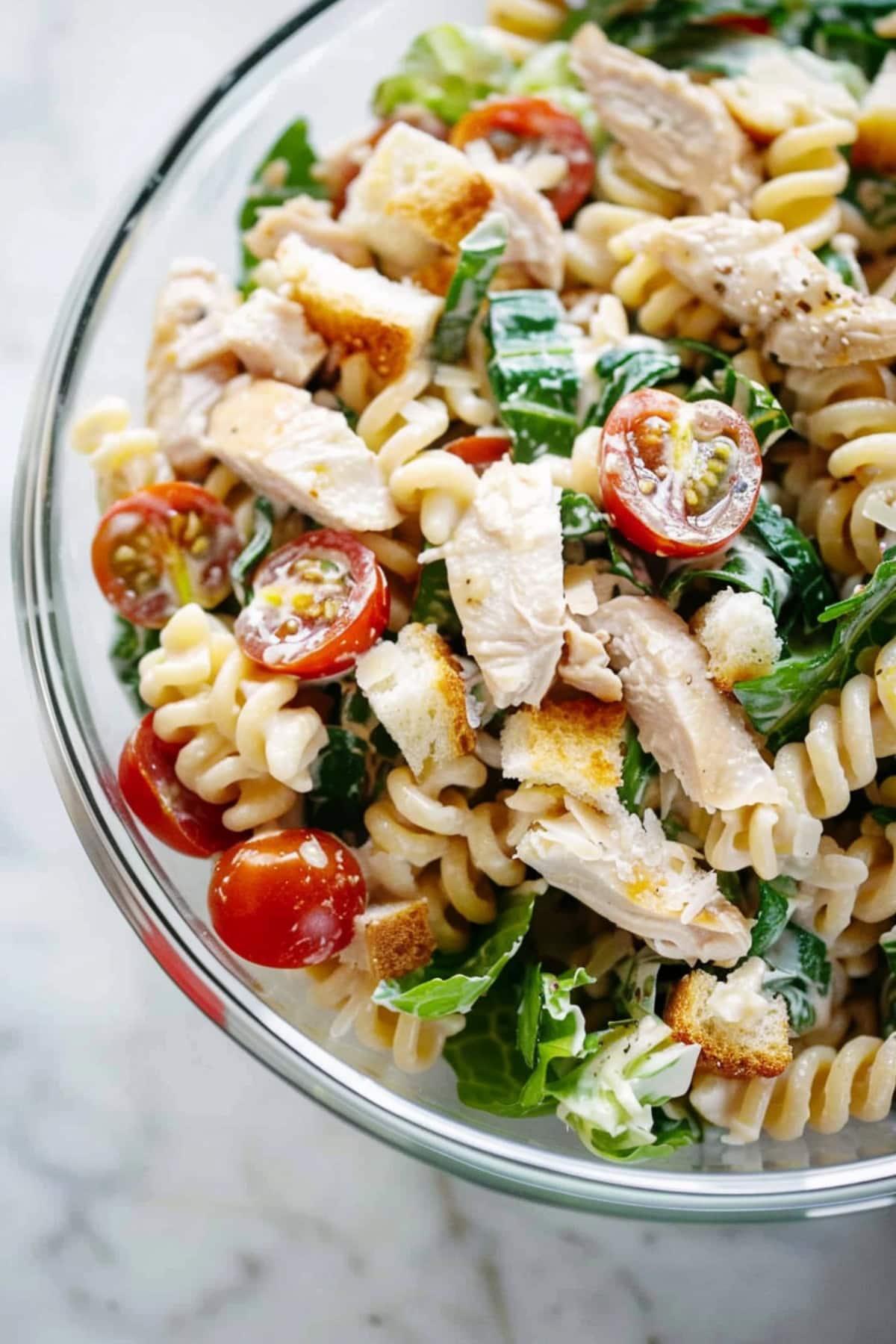 Savory and creamy rotini pasta salad with tender chicken, fresh green lettuce and cherry tomatoes in a glass bowl.