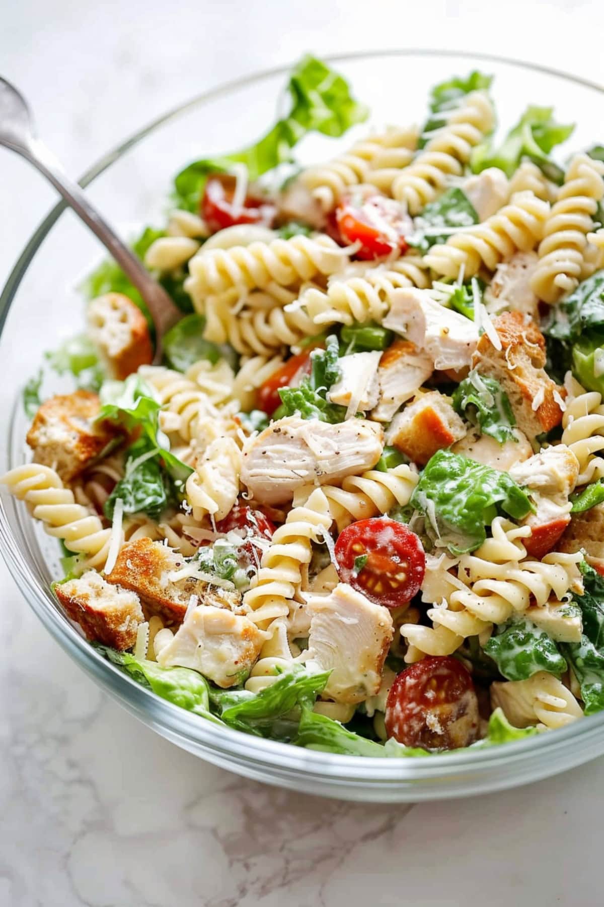 A glass bowl of chicken Caesar pasta salad with croutons, cherry tomatoes and lettuce.