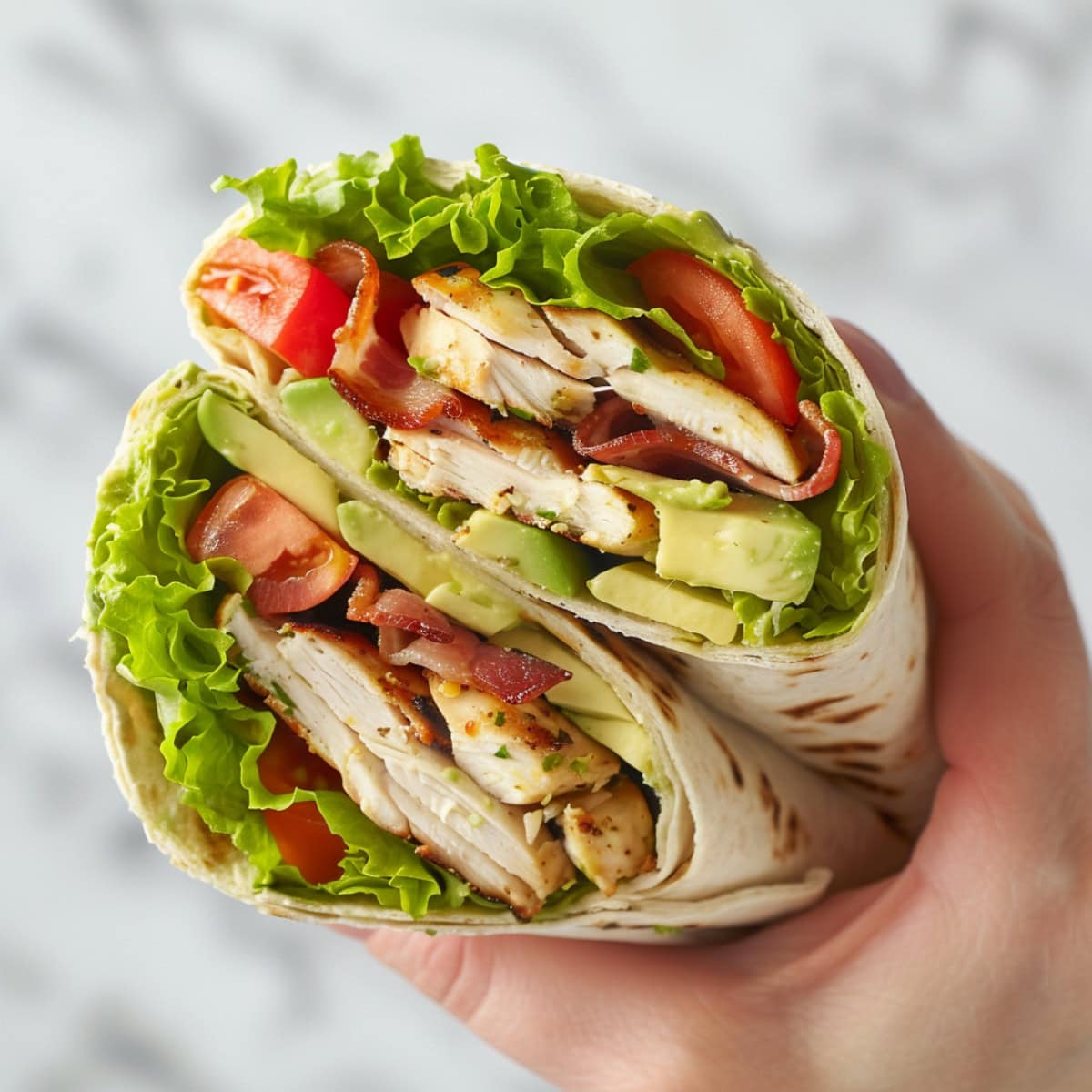 Hand holding chicken club wrap made with crisp lettuce, ripe avocado, juicy tomatoes, and crispy bacon in a soft tortilla wrap.