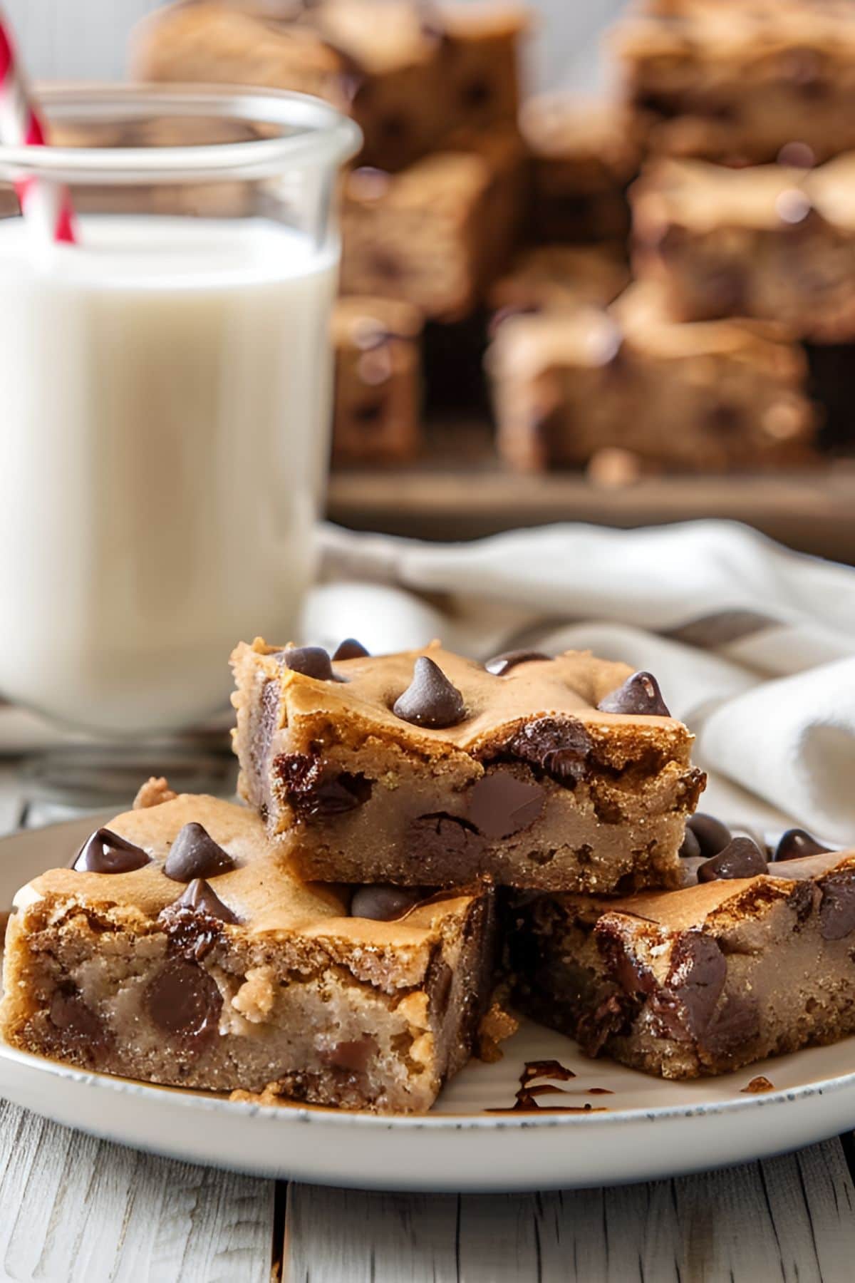 Chocolate Chip Blondie Squares on a Plate with a Glass of Milk and More Blondies in the Background