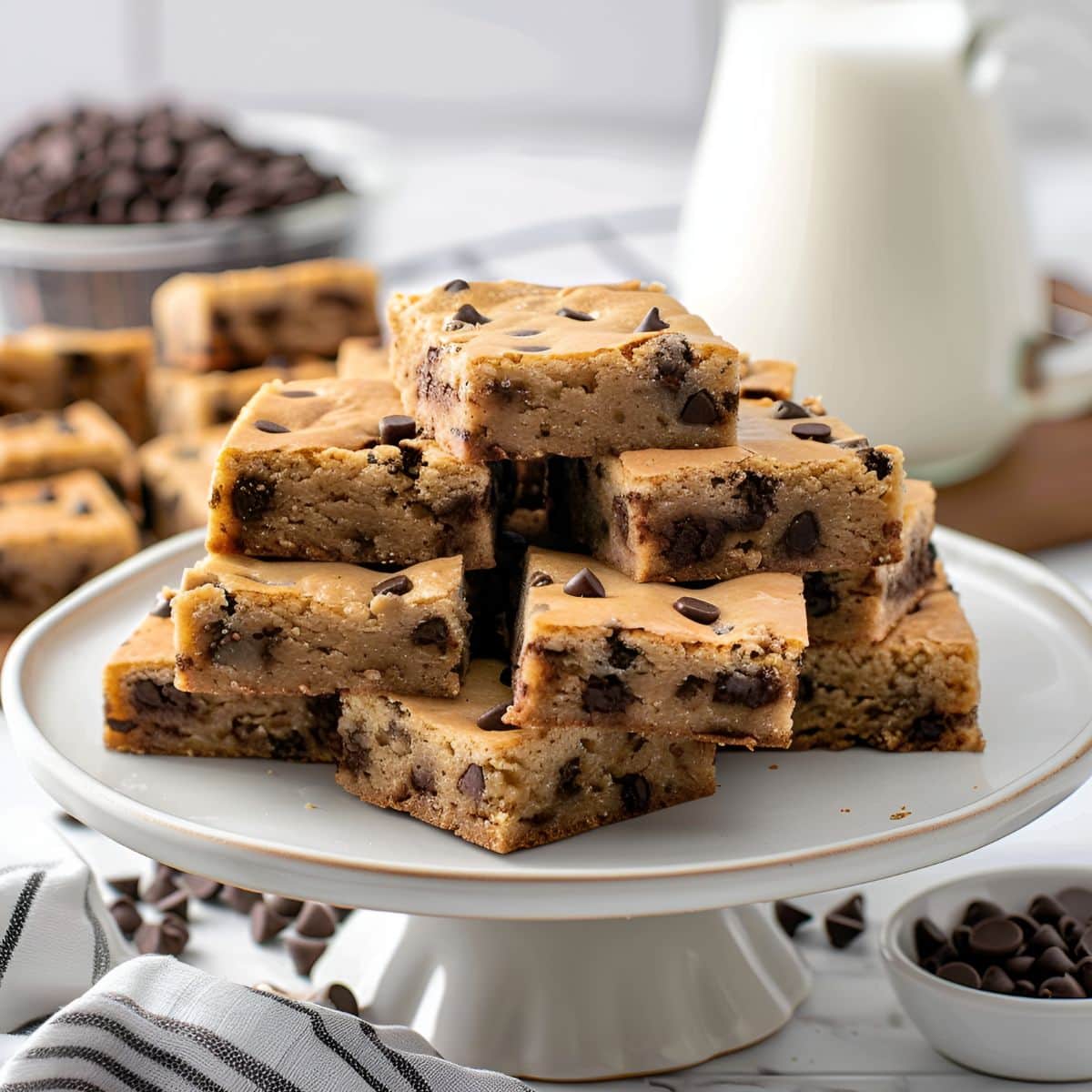 Chocolate Chip Blondie Squares Piled on a Cake Plate with More Chocolate Chip Blondies, Milk, and Chocolate Chips in the Background