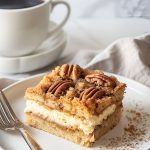 Slice of Cream Cheese Coffee Cake with Layers of Coffee Cake, Cheesecake, and Crumbly Topping with Pecans on a White Plate with a Fork and a Coffee Mug in the Background
