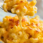 Super Close Up of Bite of Cheesy Trisha Yearwood's Crockpot Mac and Cheese on a Spoon Over a Bowl of Mac and Cheese