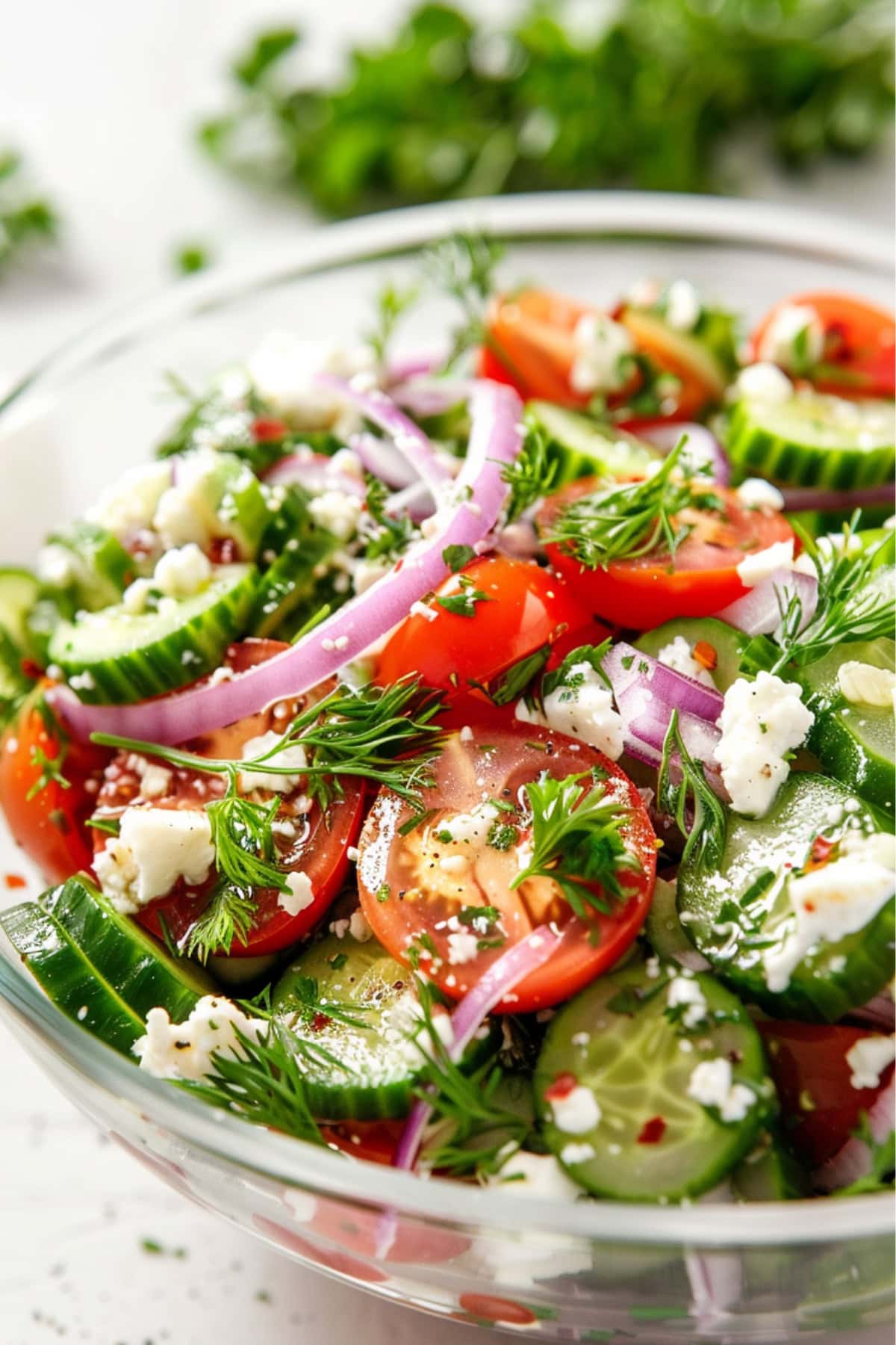 Sliced cucumbers, halved cherry tomatoes, thinly sliced red onion, feta cheese, chopped dill, and parsley in a glass bowl.