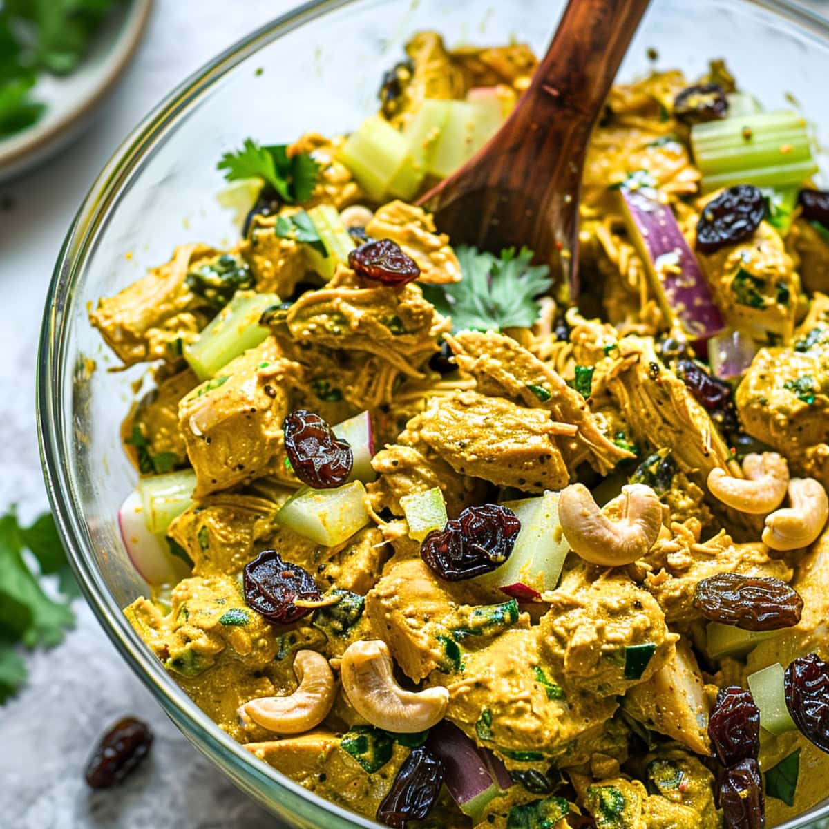 Chicken curry salad tossed with a wooden spoon on a large glass mixing bowl. 