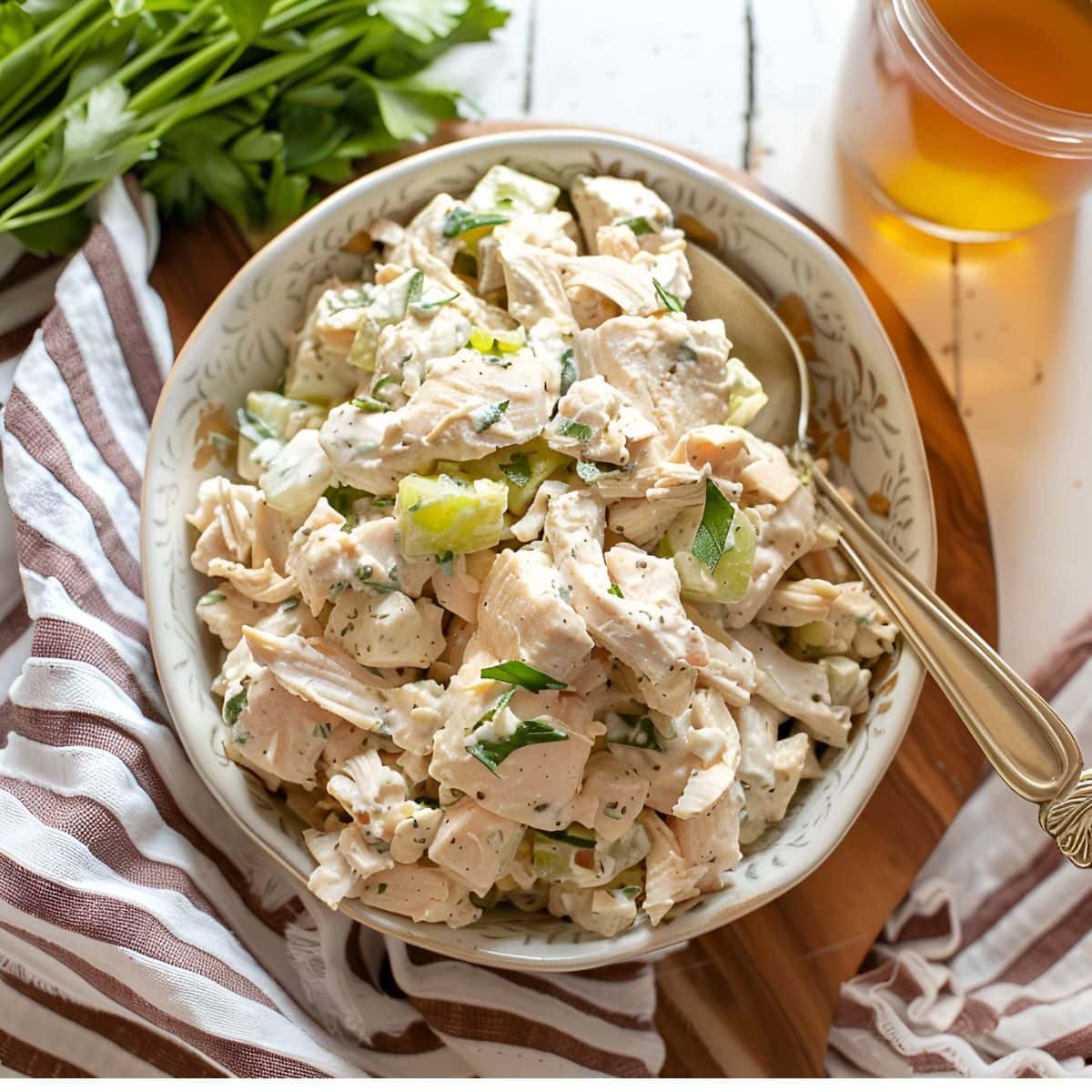 Top View of Ina Garten's Chicken Salad in a White, Patterned Bowl with a Spoon on a Wooden Cutting Board on a White Table with a Kitchen Towel