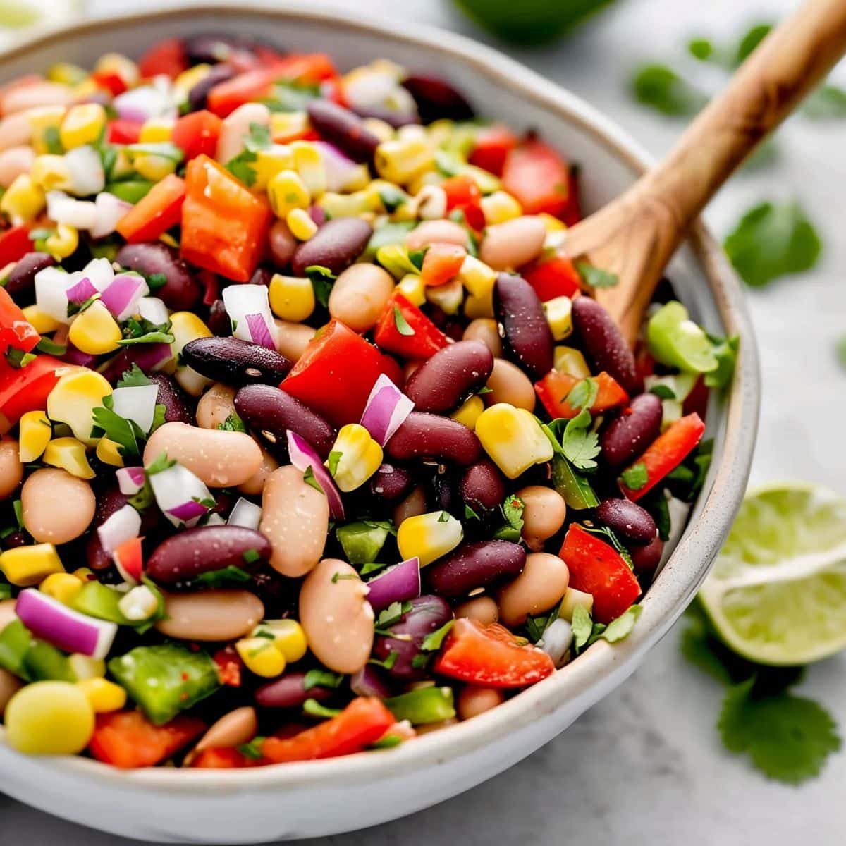 Close Up of Mexican Bean Salad in a Bowl with a Wooden Spoon, Limes, and Cilantro