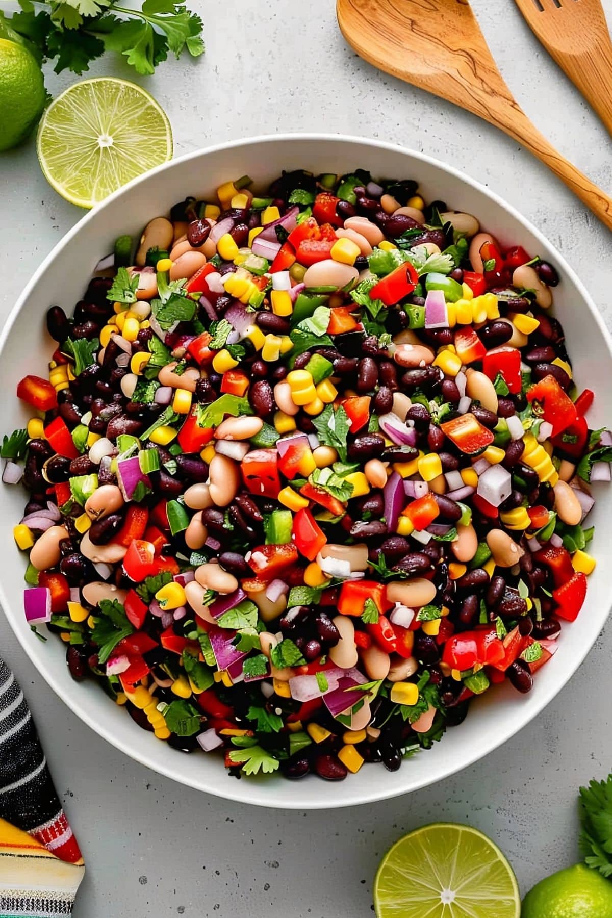 Close Up Top View of Mexican Bean Salad with Black Beans, White Beans, Peppers, Tomatoes, Corn, Red Onion, and Cilantro in a Bowl on a White Marble Table with Wooden Spoons and Limes