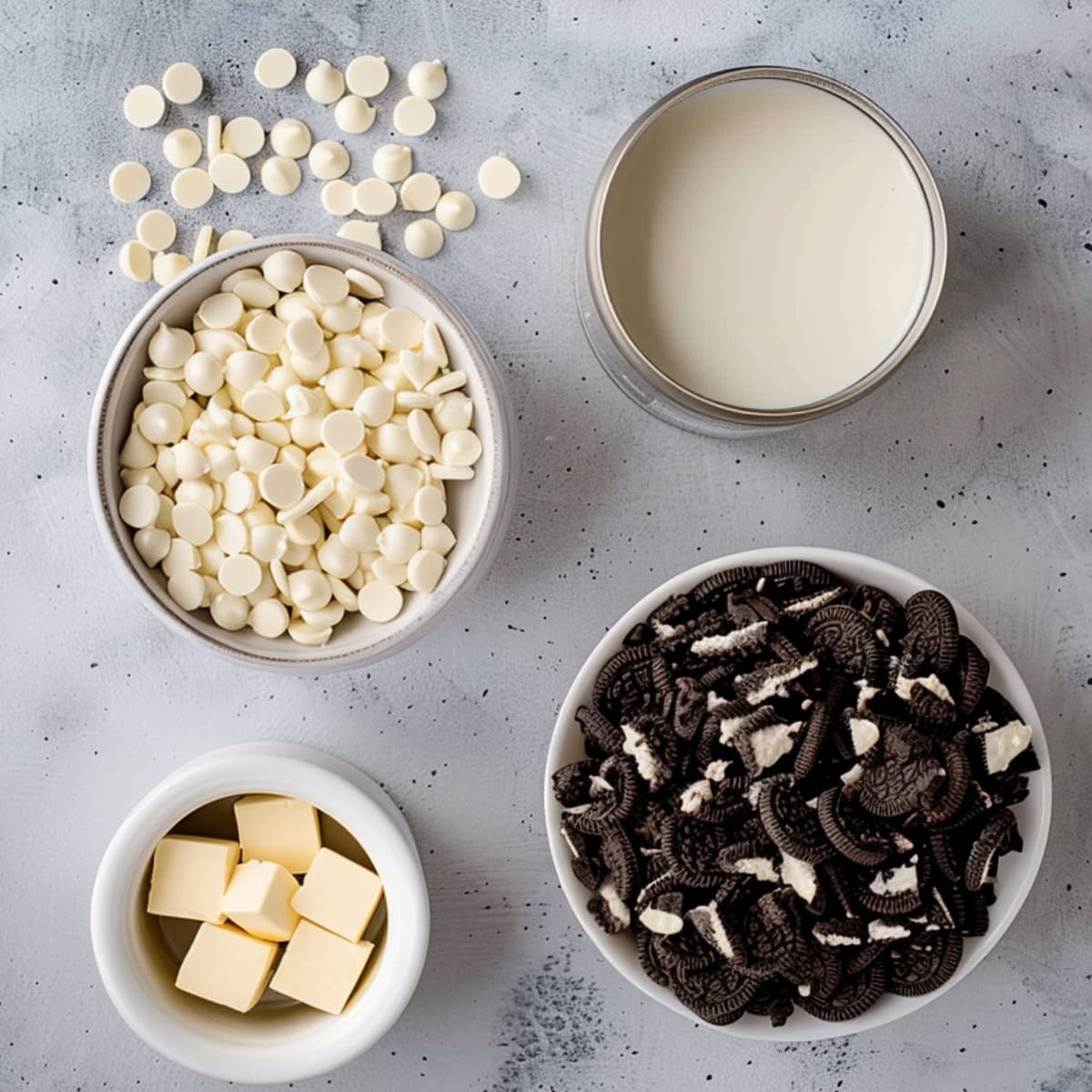 Mini white chocolate chips in a white bowl, crushed Oreos. butter and condensed mil in a can flat lay on a concrete surface.