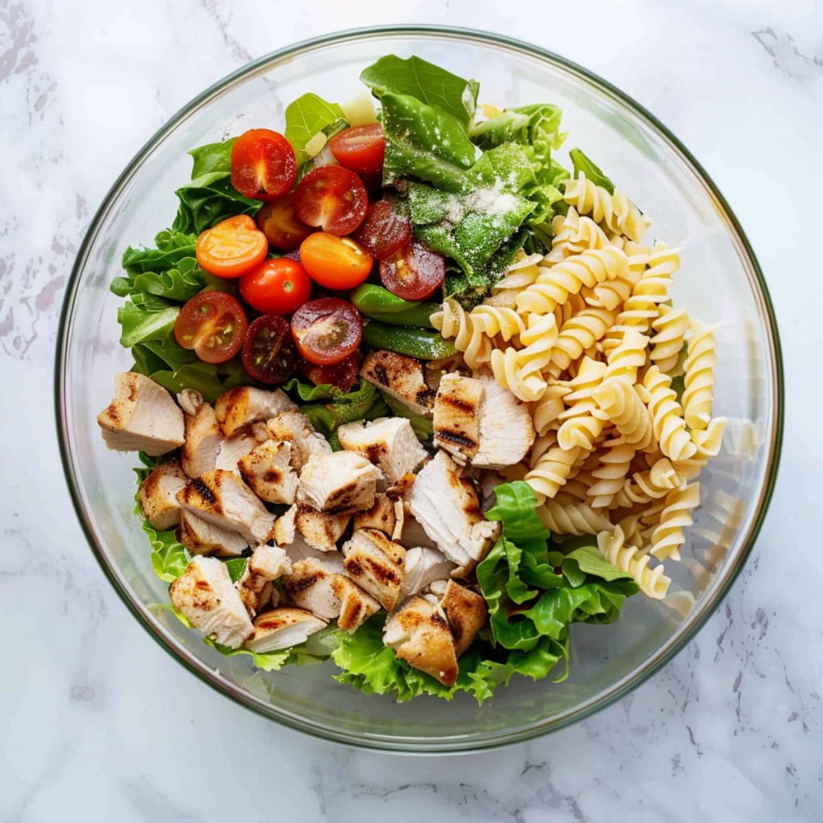 A glass bowl of grilled diced chicken, rotini pasta, cherry tomatoes and lettuce.