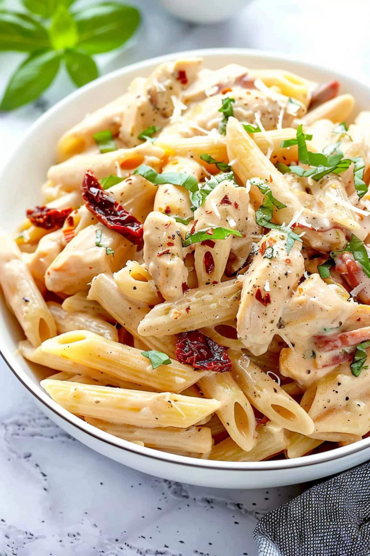 Penne pasta with heavy creamy sauce mixed with skinless chicken breasts, cut into bite-sized pieces, sun-dried tomatoes, grated Parmesan cheese, red pepper flakes, chopped baby spinach garnished with fresh basil leaves served on a white plate.